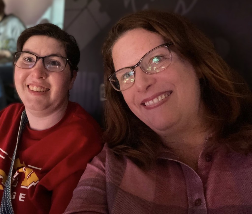 Two women wearing long sleeve shirts smiling at the camera while sitting at a indoor stadium.